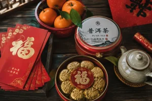 Flat-lay of Chinese New Year gifts: red envelopes, mandarin oranges, pu'er tea, and pastry tins on a wooden table
